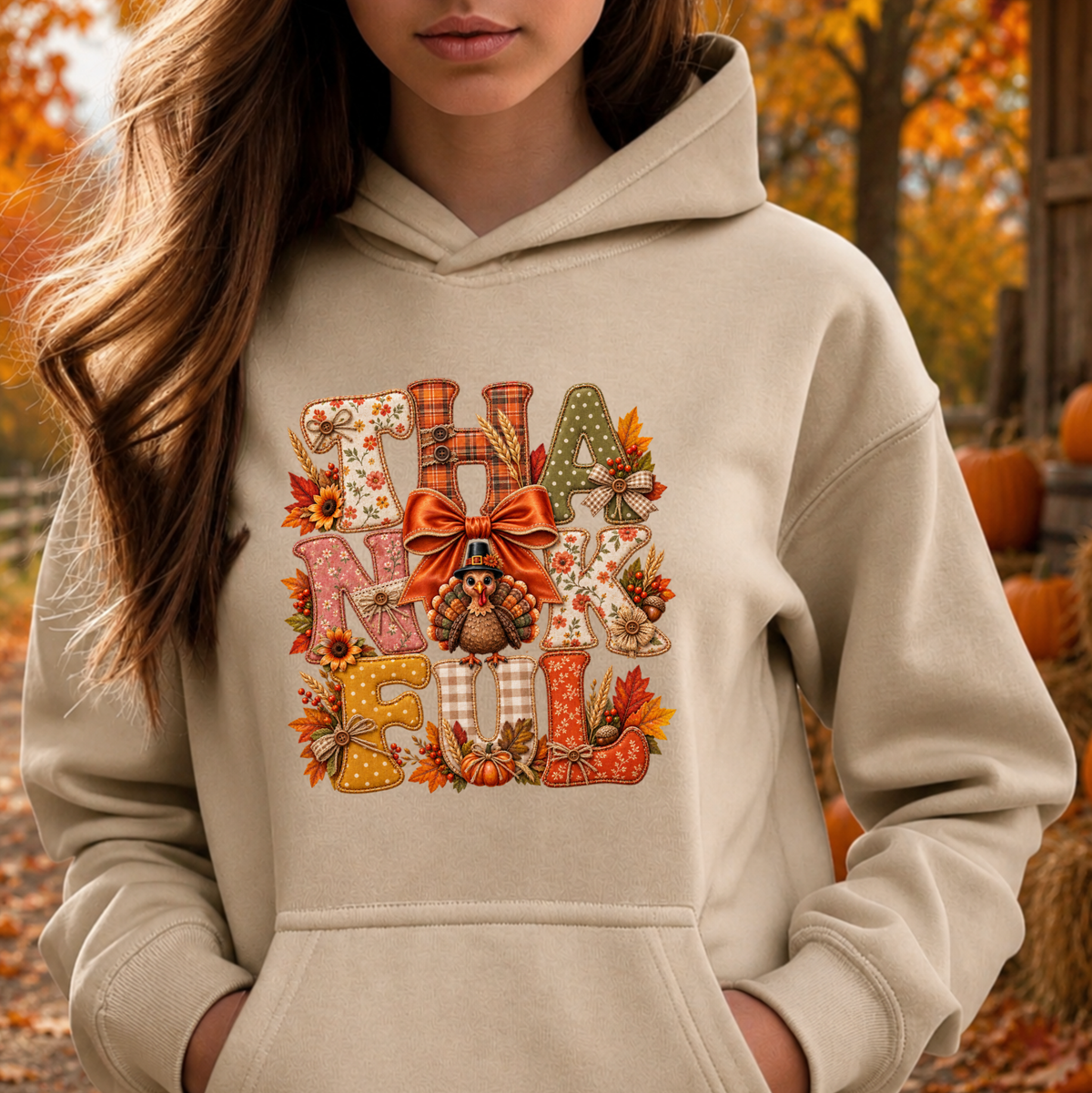 A woman wearing a beige hoodie with a large autumn-themed 'Thankful Fall' graphic on the front, set against an outdoor fall background with pumpkins and orange foliage.