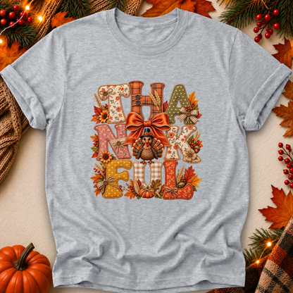 A heather gray short-sleeve t-shirt with a large 'THANKsgiving' graphic print featuring autumn-themed letters, a turkey, and a bow. The shirt is displayed in a flat-lay setting decorated with fall leaves, pine branches, a pumpkin, and string lights.