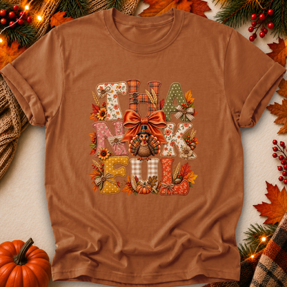 A brown short-sleeve crewneck t-shirt with a colorful 'THANKFUL' graphic featuring autumn motifs like pumpkins, leaves, and a bow, presented in a flat-lay with seasonal props.