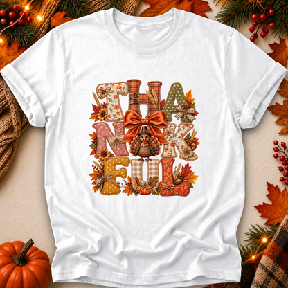 A white short-sleeve crewneck t-shirt with a large 'THANK FELL' graphic printed on the front, styled in a flat-lay autumn setting with pumpkins, pine branches, and fall leaves.