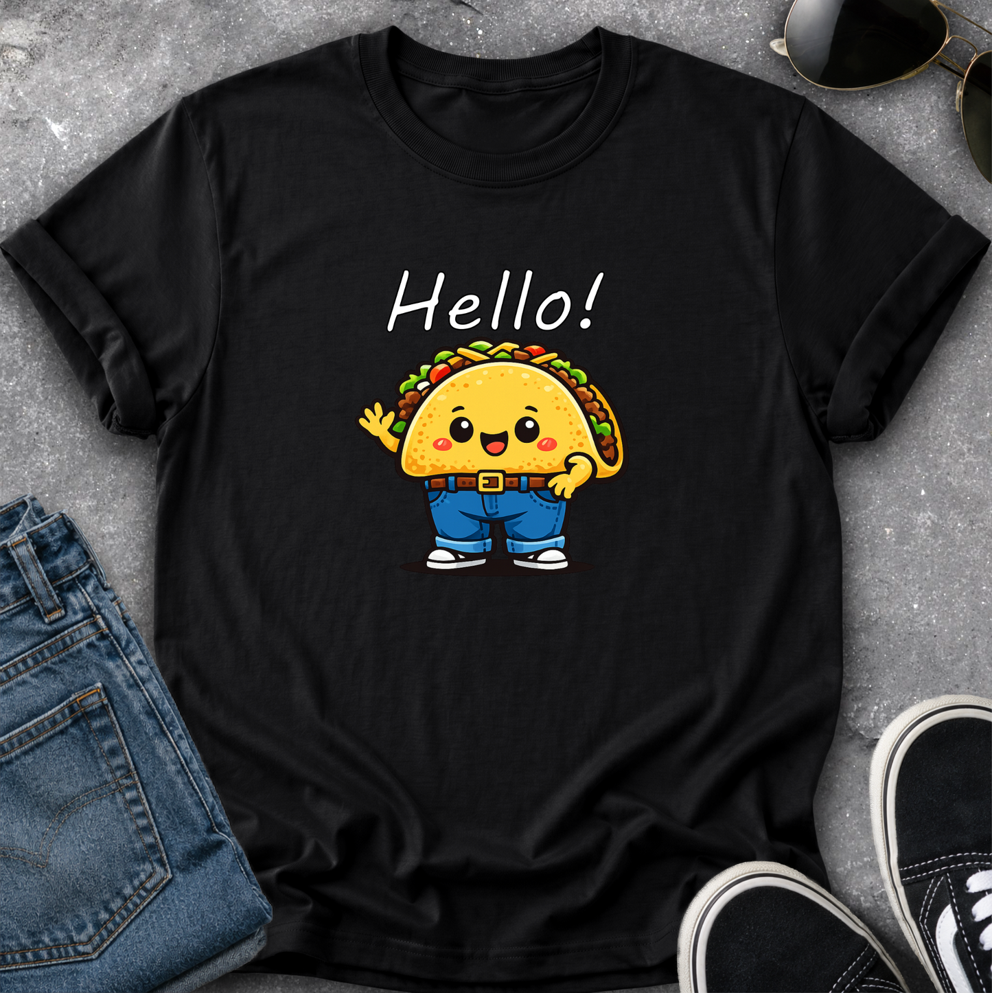 A black short-sleeve graphic T-shirt laid flat on a gray textured surface, styled with blue jeans, sunglasses, and black sneakers. The shirt features a cartoon taco character wearing blue pants and white sneakers, with the word 'Hello!' printed above in white.