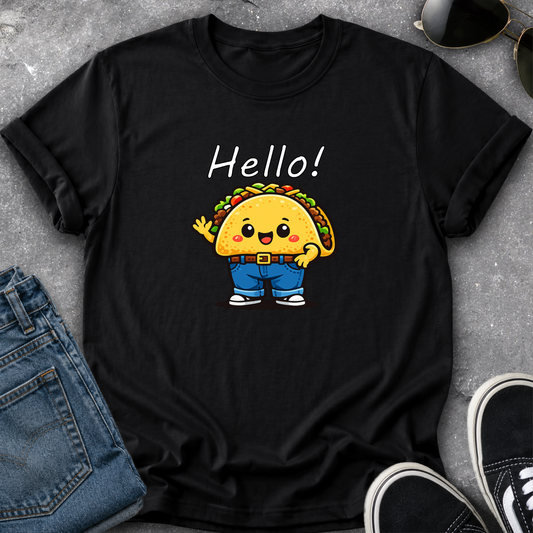 A black short-sleeve graphic T-shirt laid flat on a gray textured surface, styled with blue jeans, sunglasses, and black sneakers. The shirt features a cartoon taco character wearing blue pants and white sneakers, with the word 'Hello!' printed above in white.