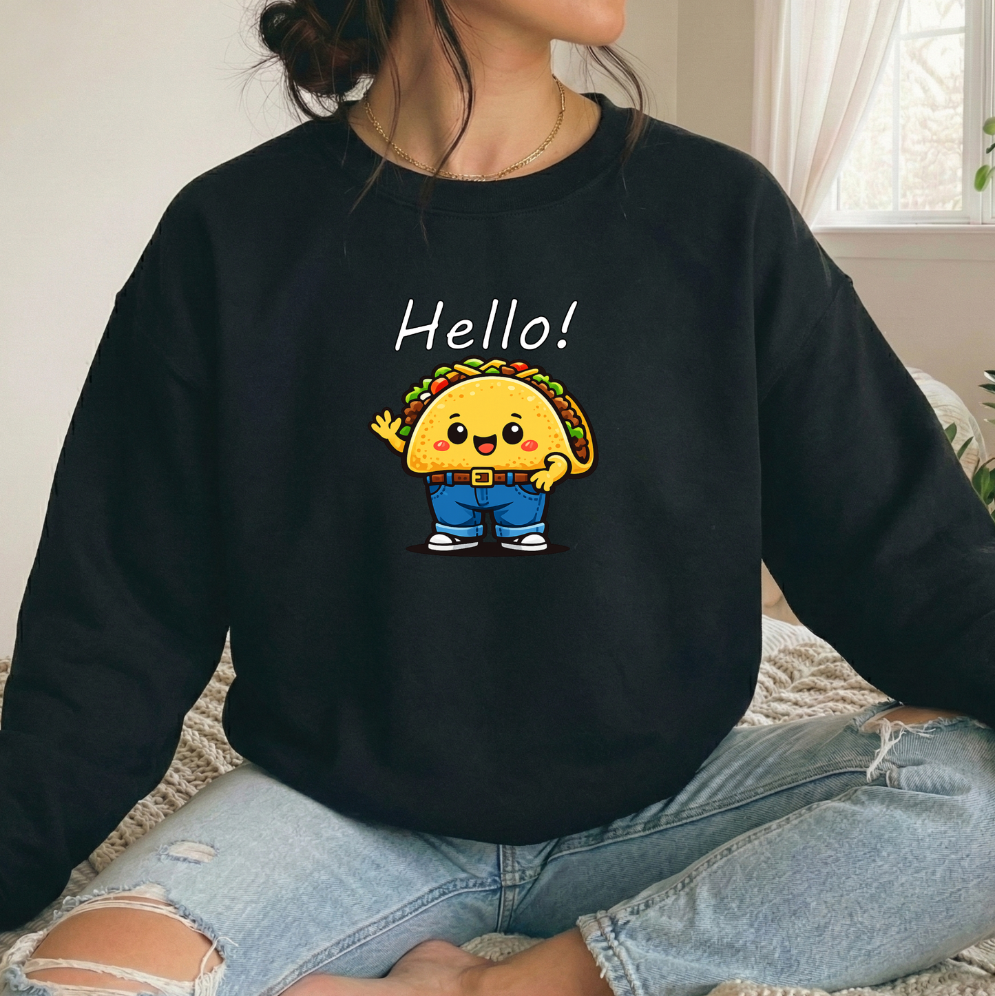 A person wearing a black crewneck sweatshirt with a cartoon taco graphic and the word 'Hello!' printed on the front, paired with light blue ripped jeans in a bright indoor setting.
