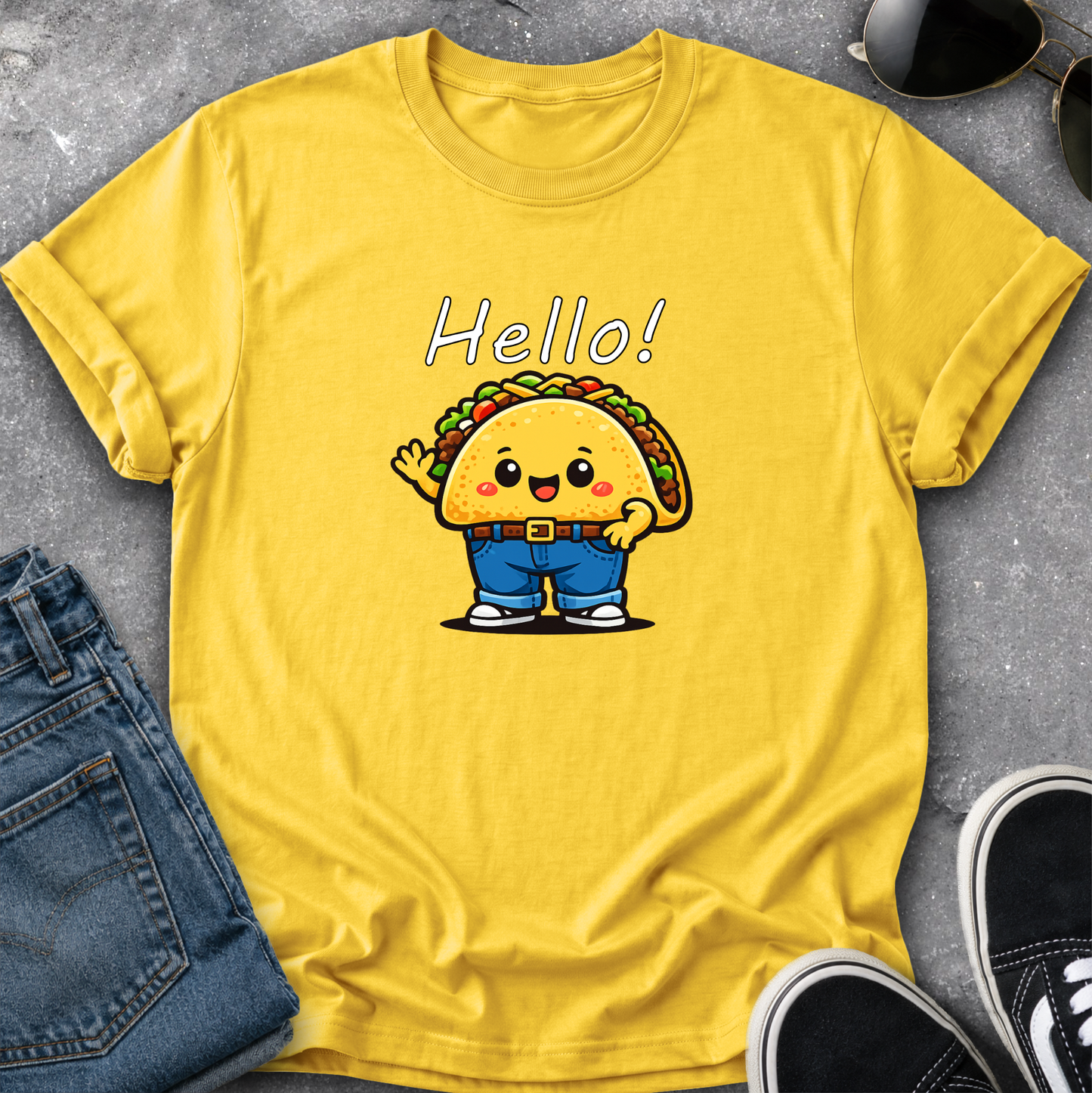 A yellow short-sleeve t-shirt with a cartoon taco character and the word 'Hello!' printed on the front, styled in a flat lay with blue jeans, sunglasses, and black sneakers on a gray textured surface.