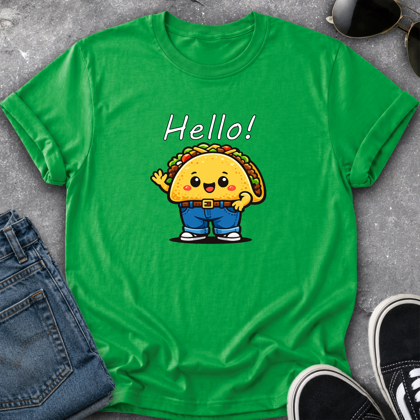 A bright green short-sleeve t-shirt laid flat on a gray textured surface, featuring a cartoon taco character wearing blue pants and the word 'Hello!' printed above it. Blue jeans, sunglasses, and black sneakers are styled around the shirt.