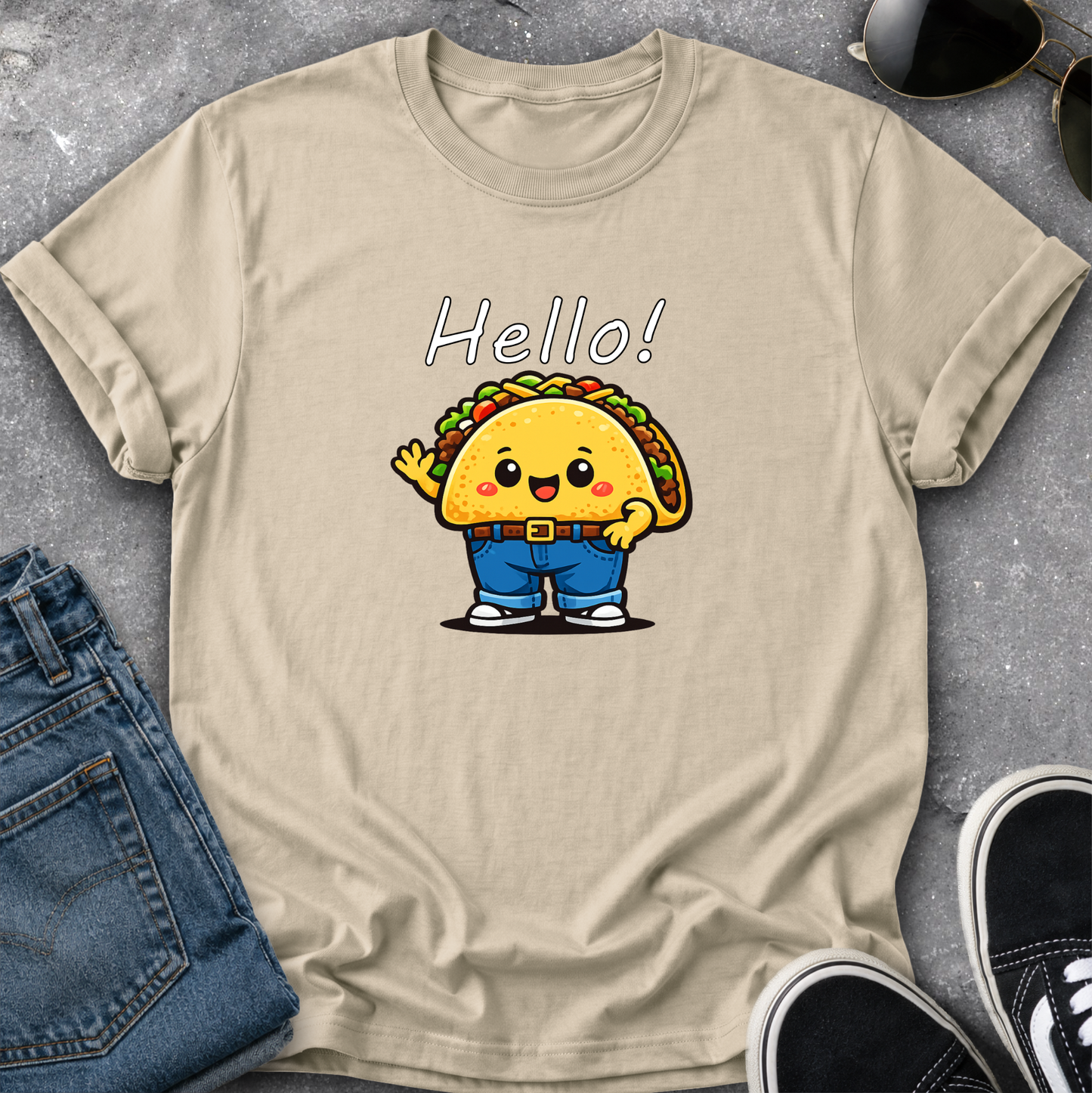 A beige graphic t-shirt laid flat on a gray textured surface, styled with blue jeans, sunglasses, and black sneakers. The t-shirt features a cartoon taco character wearing blue pants and waving, with the word 'Hello!' printed above it.