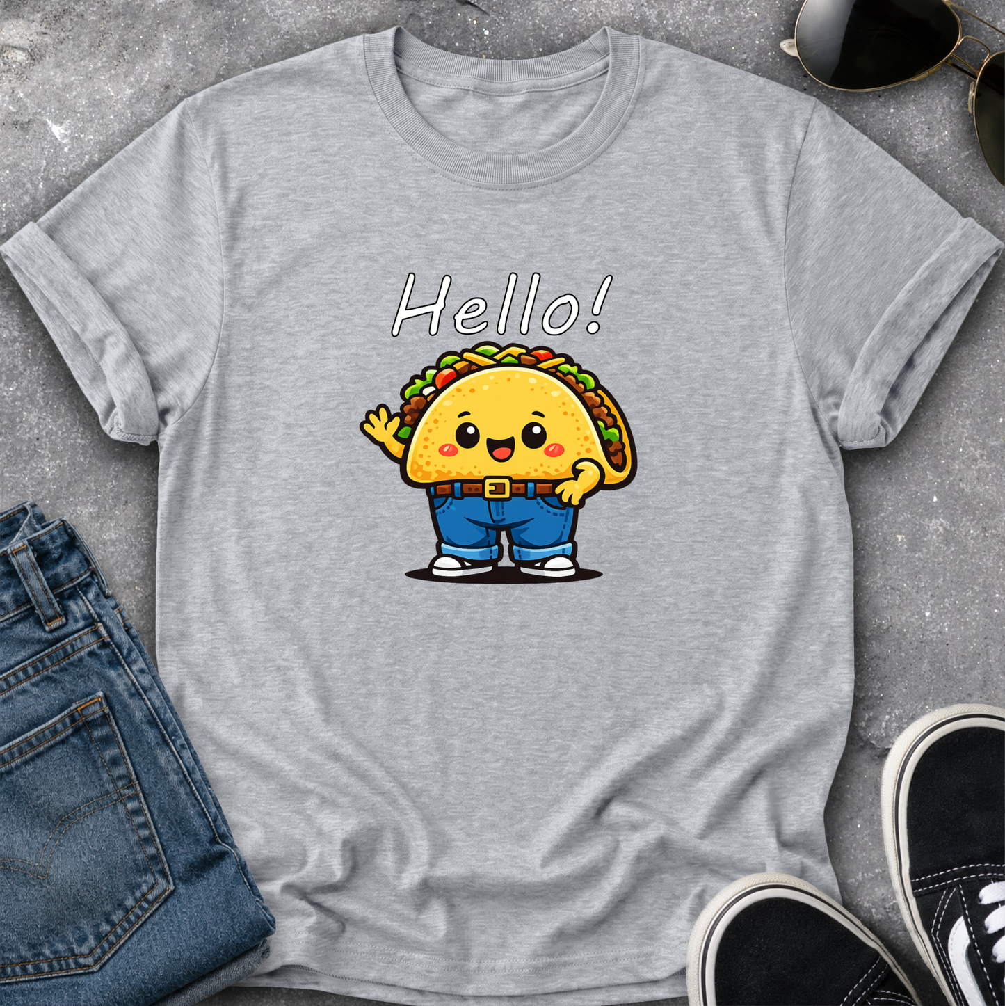 A heather gray short-sleeve t-shirt with a cute cartoon taco character wearing blue pants and the word 'Hello!' printed above it. The shirt is styled in a flat lay with blue jeans, sunglasses, and black sneakers on a gray textured surface.