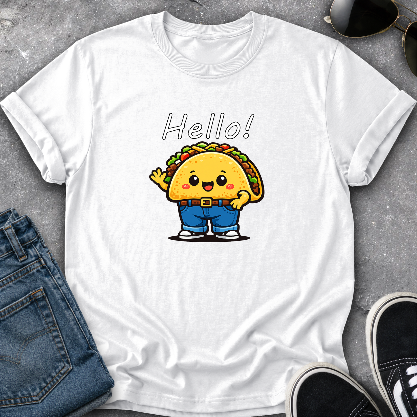 A white short-sleeve crewneck t-shirt laid flat on a gray textured surface, featuring a cute cartoon taco character wearing blue pants and waving, with the word 'Hello!' printed above. Blue jeans, sunglasses, and black sneakers are styled around the shirt.