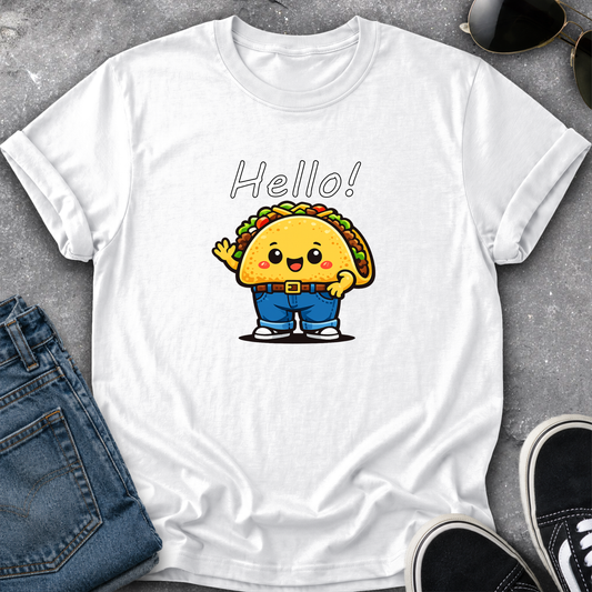 A white short-sleeve crewneck t-shirt laid flat on a gray textured surface, featuring a cute cartoon taco character wearing blue pants and waving, with the word 'Hello!' printed above. Blue jeans, sunglasses, and black sneakers are styled around the shirt.