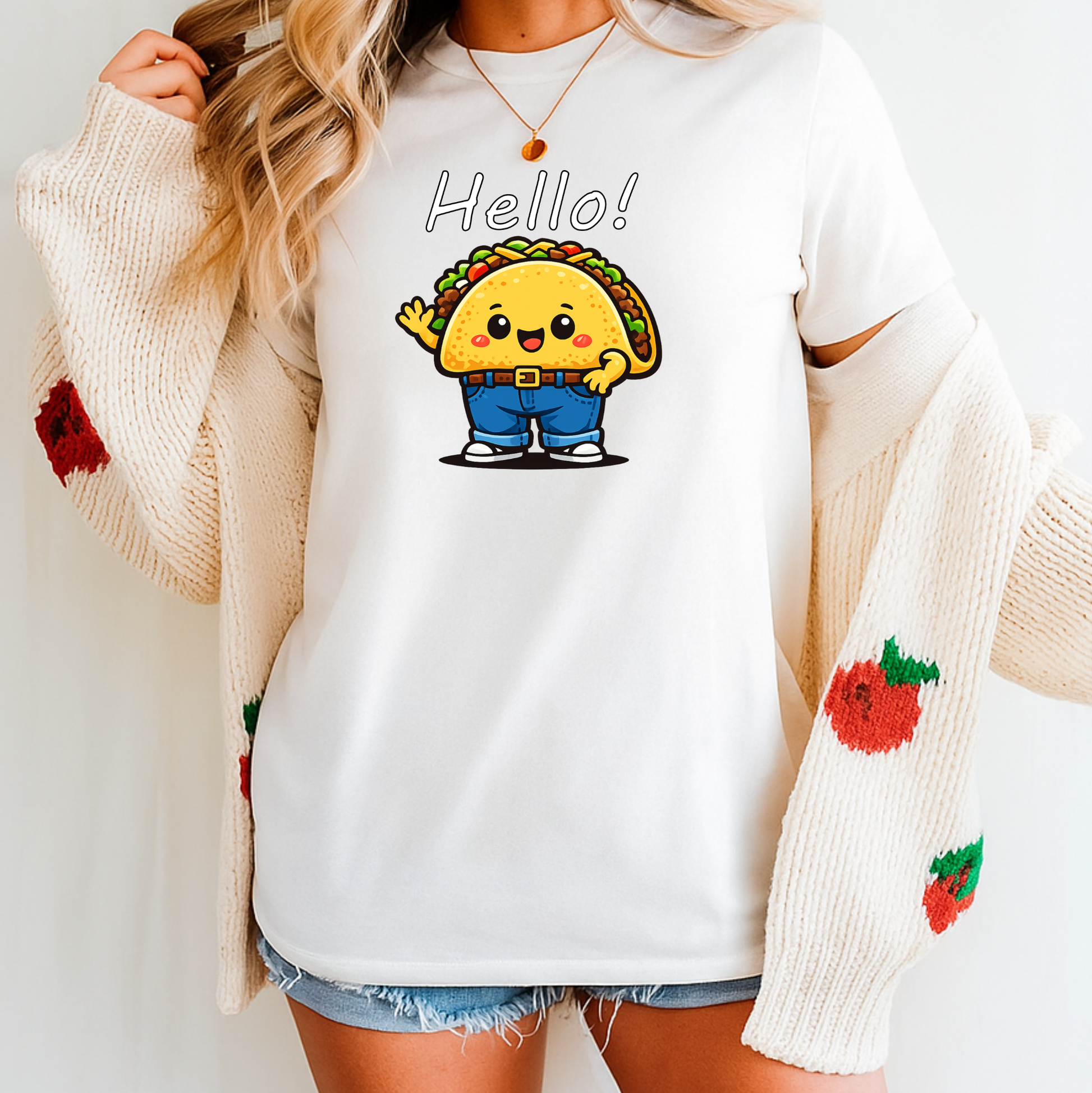 A woman wearing a white crewneck t-shirt with a cute cartoon taco graphic and the word 'Hello!' printed on the chest, styled with an open cream knit cardigan featuring red strawberry details.