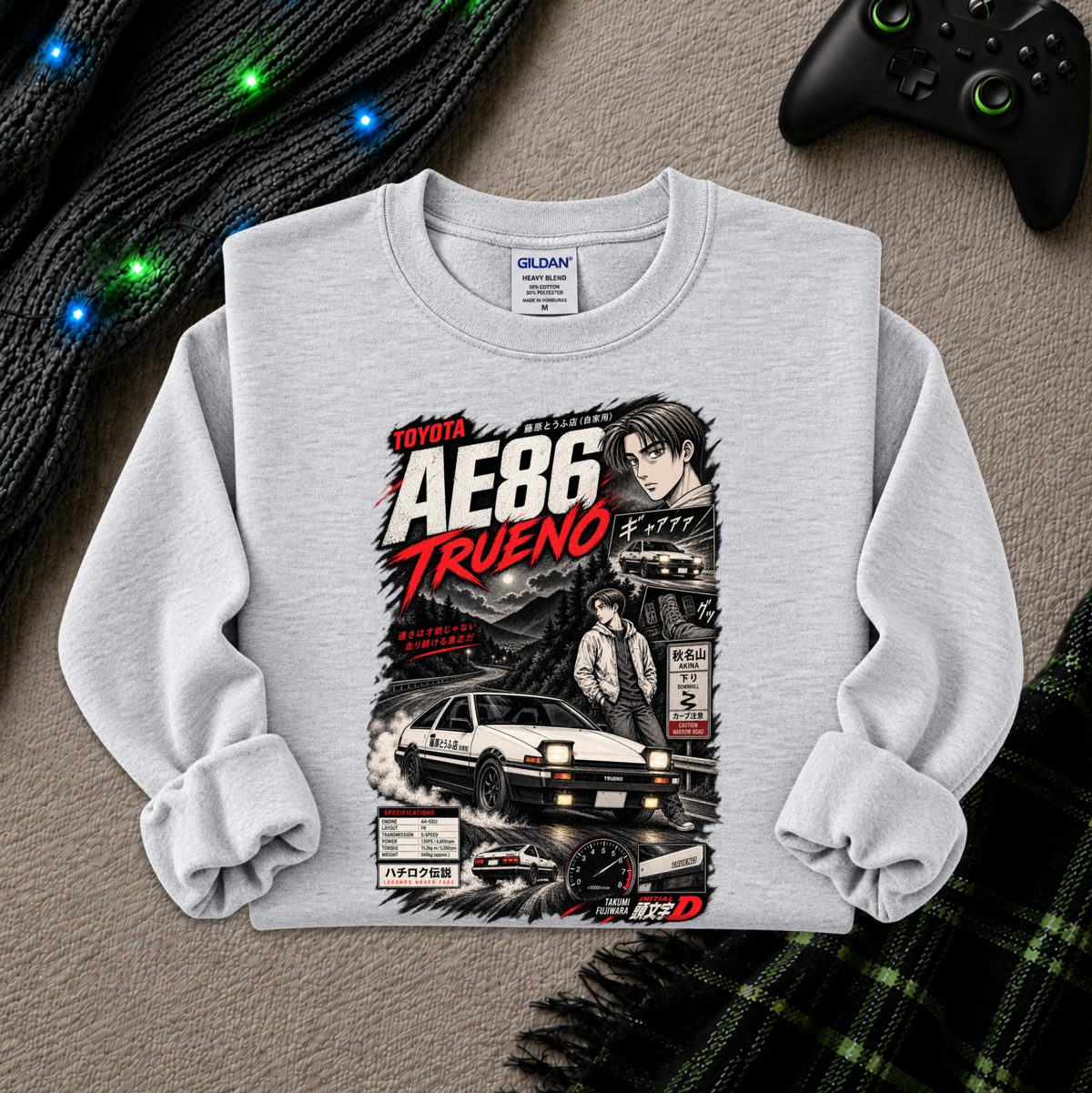 A light gray long-sleeve crewneck sweatshirt with a detailed graphic print of the Toyota AE86 Trueno from the movie Initial D, styled flat on a textured gray surface with a black controller, a knit sweater, and a plaid blanket.