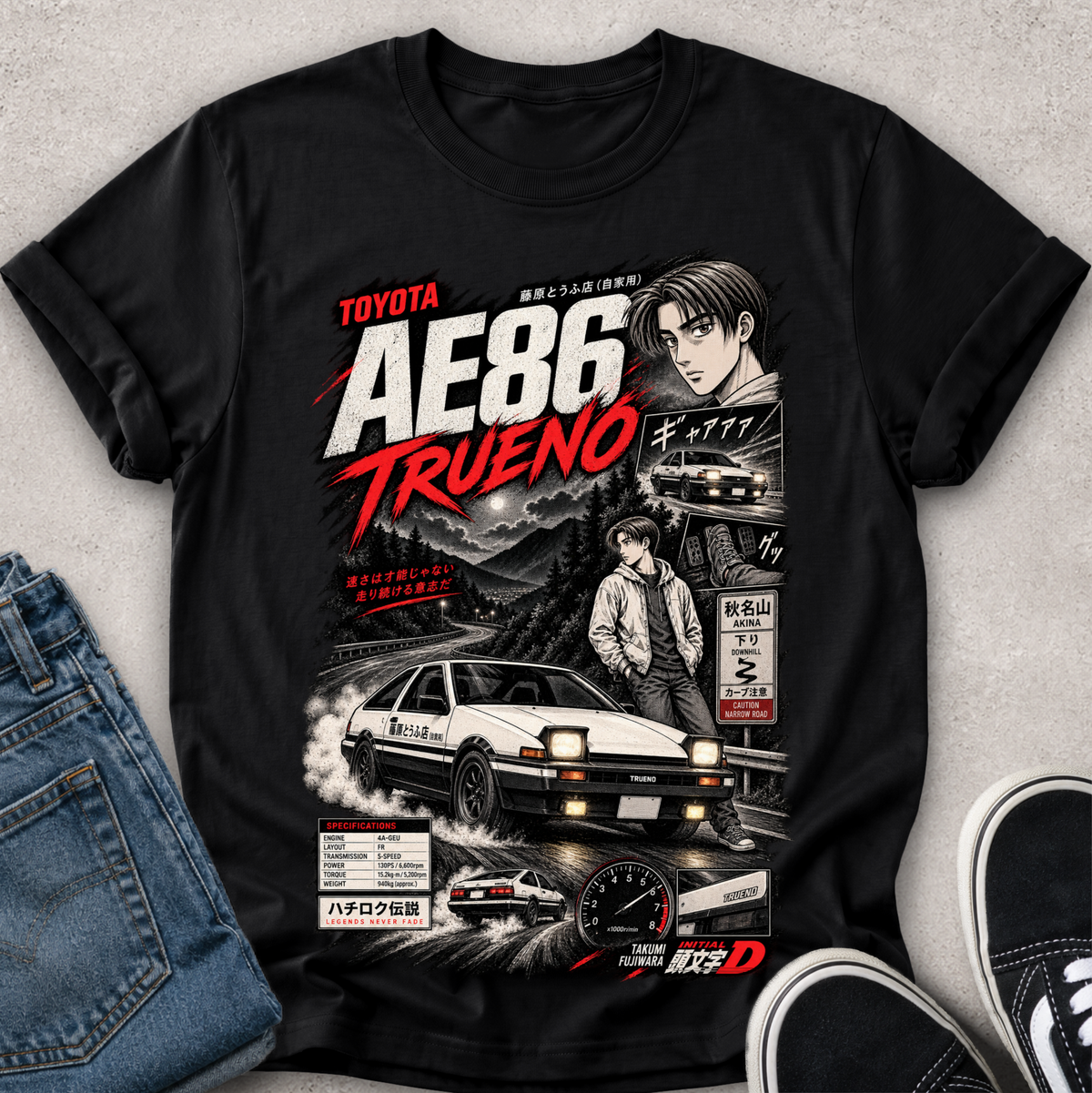 A black graphic t-shirt featuring a detailed print of the Toyota AE86 Trueno from the movie Initial D, styled in a flat-lay with blue jeans and black sneakers on a textured gray background.