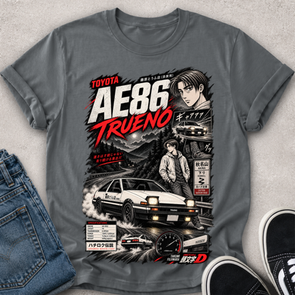 A gray graphic t-shirt featuring a detailed print of the Toyota AE86 Trueno from the movie Initial D, styled in a flat-lay with blue jeans and black sneakers on a textured gray background.