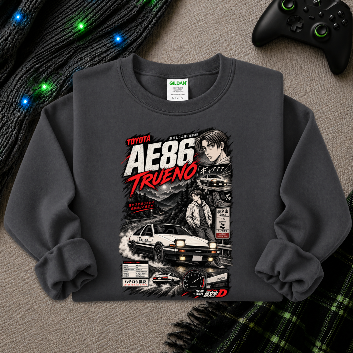 A dark gray crewneck sweatshirt featuring a detailed graphic print of the Toyota AE86 Trueno from the movie Initial D, styled in a flat-lay arrangement with a black knit sweater, glowing fairy lights, a black video game controller, and a green plaid blanket on a gray textured surface.