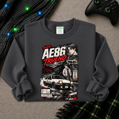 A dark gray crewneck sweatshirt featuring a detailed graphic print of the Toyota AE86 Trueno from the movie Initial D, styled in a flat-lay arrangement with a black knit sweater, glowing fairy lights, a black video game controller, and a green plaid blanket on a gray textured surface.
