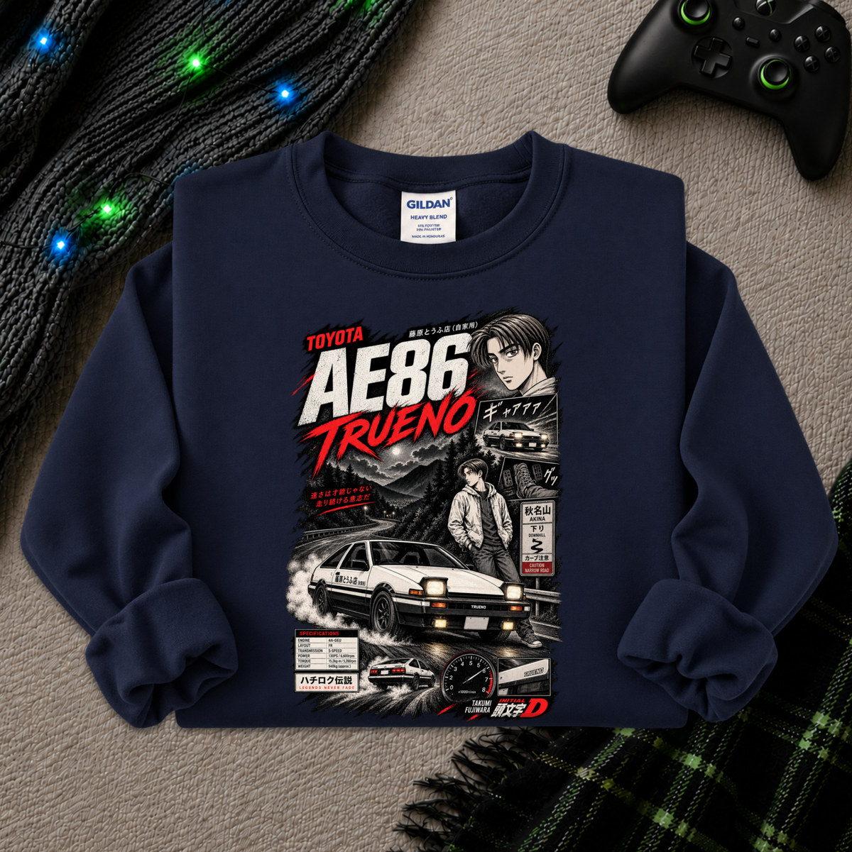 A navy blue Gildan sweatshirt featuring a detailed graphic print of the Toyota AE86 Trueno, styled in a flat-lay arrangement with a black knit sweater, glowing fairy lights, a black video game controller, and a green plaid blanket on a gray textured background.