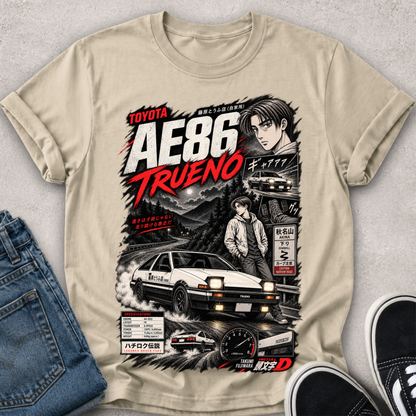 A beige graphic t-shirt featuring a detailed design inspired by the movie Initial D 'AE86 Trueno', styled in a flat-lay with blue jeans and black sneakers on a gray textured background.