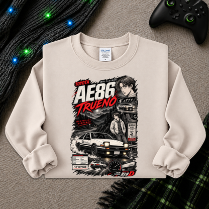 A beige long-sleeve crewneck sweatshirt featuring a detailed graphic print of the Toyota AE86 Trueno from the anime Initial D. The sweatshirt is styled flat on a gray textured surface with a black knit sweater, a black video game controller, and a green plaid blanket.