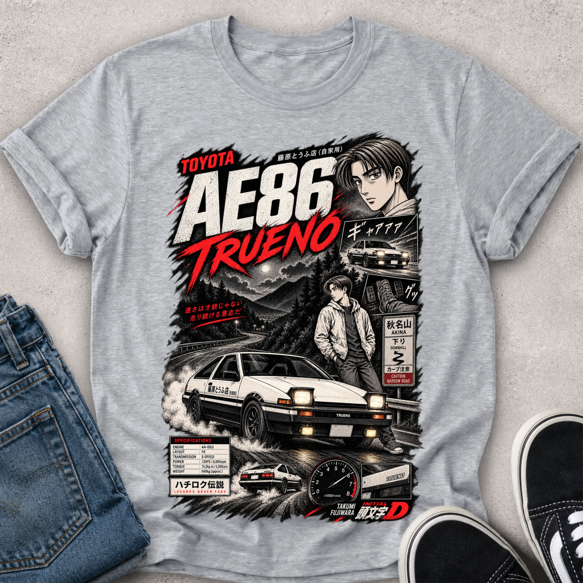 A heather gray short-sleeve graphic T-shirt laid flat on a light textured surface, styled with blue jeans on the left and black sneakers on the right. The shirt features a large front print with the words 'TOYOTA AE86 TRUENO,' a car illustration, a character portrait, and Japanese text.