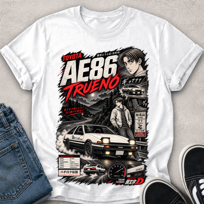 A white short-sleeve graphic T-shirt laid flat on a light textured surface, styled with blue jeans on the left and black sneakers on the right. The shirt features a large front print inspired by the Toyota AE86 Trueno, with illustrated characters, a car, road signs, and bold red and black text.