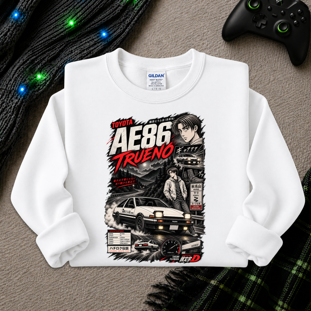 A white long-sleeve crewneck sweatshirt with a detailed graphic print of the Toyota AE86 Trueno, styled flat on a gray textured background with a game controller and festive lights nearby.