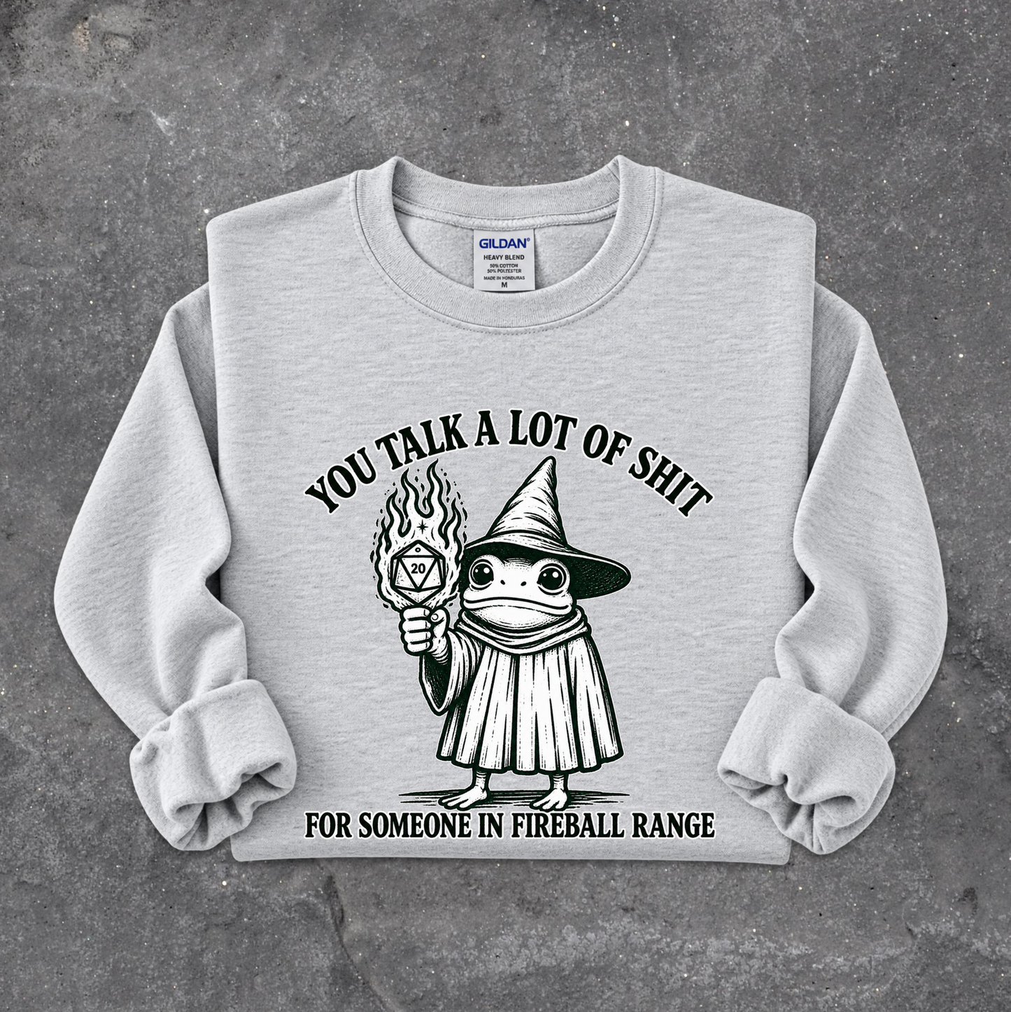 A light gray crewneck sweatshirt with a black graphic of a cartoon witch holding a fireball, with the text 'YOU TALK A LOT OF SHIT FOR SOMEONE IN FIREBALL RANGE'. The sweatshirt is laid flat on a textured gray concrete surface.