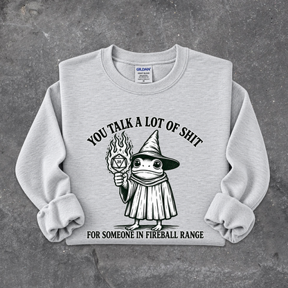 A light gray crewneck sweatshirt with a black graphic of a cartoon witch holding a fireball, with the text 'YOU TALK A LOT OF SHIT FOR SOMEONE IN FIREBALL RANGE'. The sweatshirt is laid flat on a textured gray concrete surface.