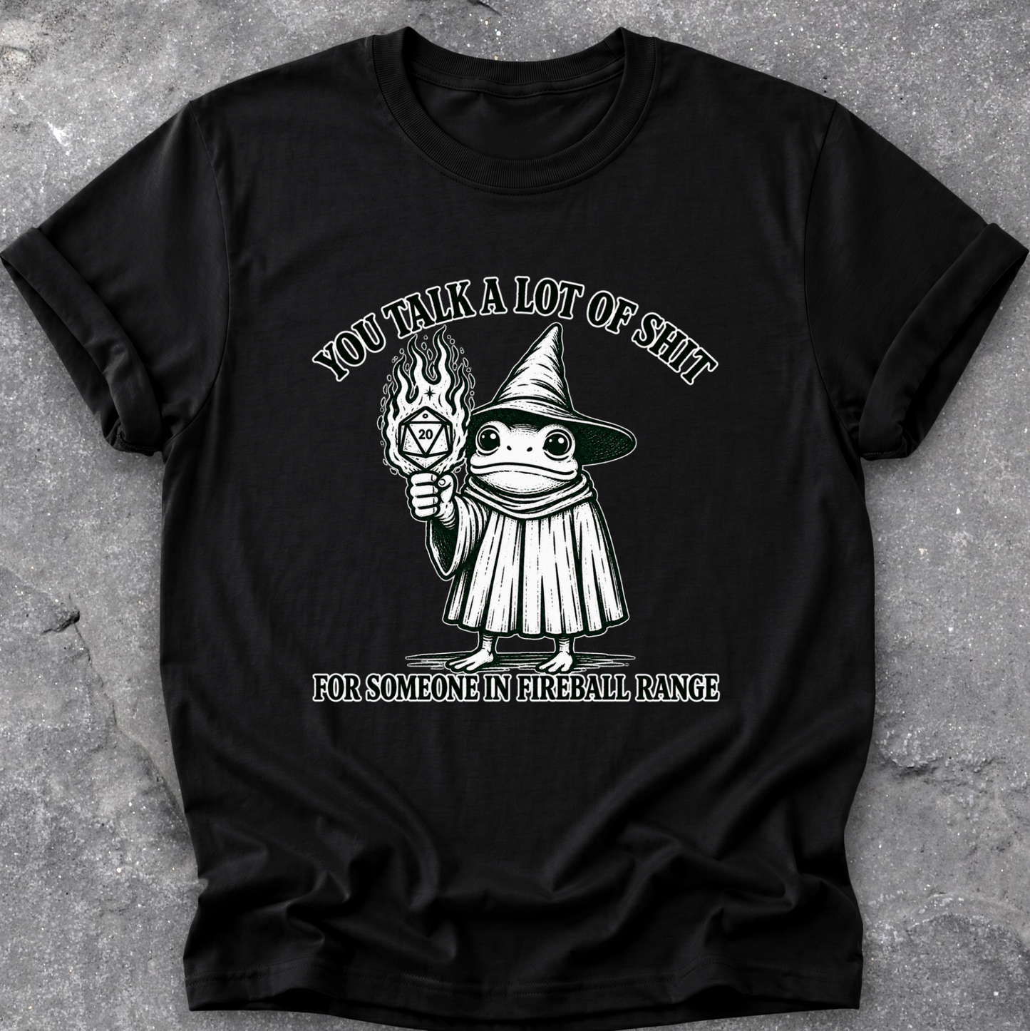 A black short-sleeve graphic t-shirt laid flat on a textured gray concrete surface. The graphic features a cartoon frog dressed as a witch holding a flaming torch, with the text 'YOU TALK A LOT OF SHIT' above and 'FOR SOMEONE IN FIREBALL RANGE' below.