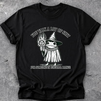 A black short-sleeve graphic t-shirt laid flat on a textured gray concrete surface. The graphic features a cartoon frog dressed as a witch holding a flaming torch, with the text 'YOU TALK A LOT OF SHIT' above and 'FOR SOMEONE IN FIREBALL RANGE' below.