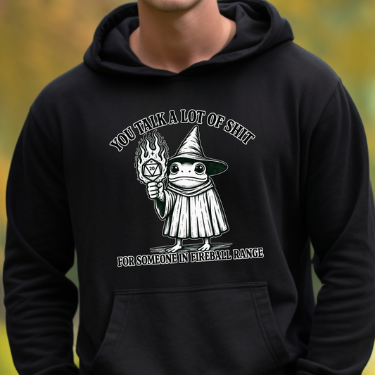 A person wearing a black pullover hoodie with a white graphic print on the chest. The graphic shows a cartoon wizard holding a flame and text that reads 'YOU TALK A LOT OF SHIT FOR SOMEONE IN FIREBALL RANGE'.