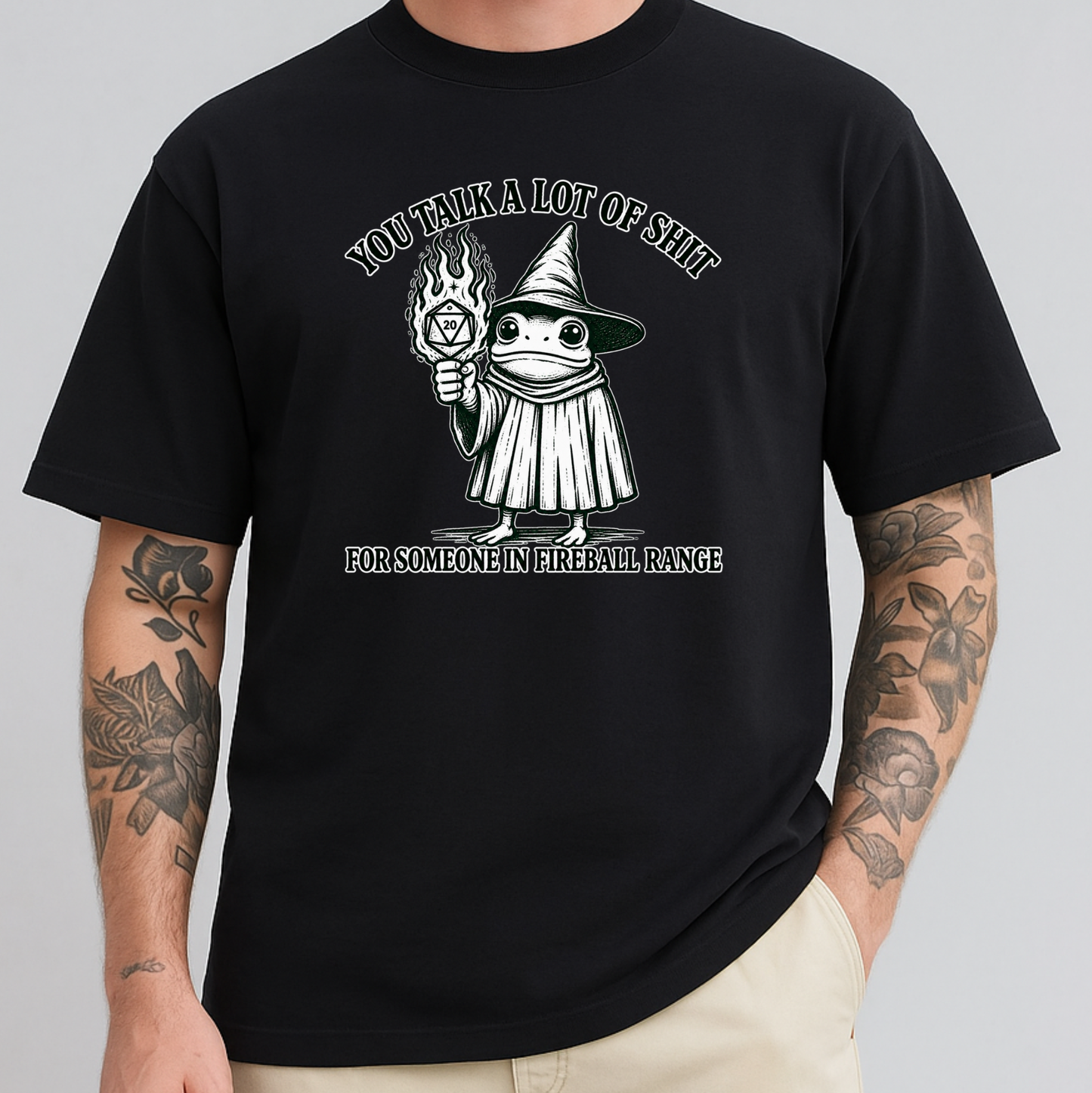A man wearing a black graphic t-shirt with a white and gray illustration of a wizard holding a flaming orb, with the text 'YOU TALK A LOT OF SHIT' above and 'FOR SOMEONE IN FIREBALL RANGE' below. The model has tattooed arms and is wearing beige pants, set against a solid gray background.