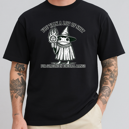 A man wearing a black graphic t-shirt with a white and gray illustration of a wizard holding a flaming orb, with the text 'YOU TALK A LOT OF SHIT' above and 'FOR SOMEONE IN FIREBALL RANGE' below. The model has tattooed arms and is wearing beige pants, set against a solid gray background.