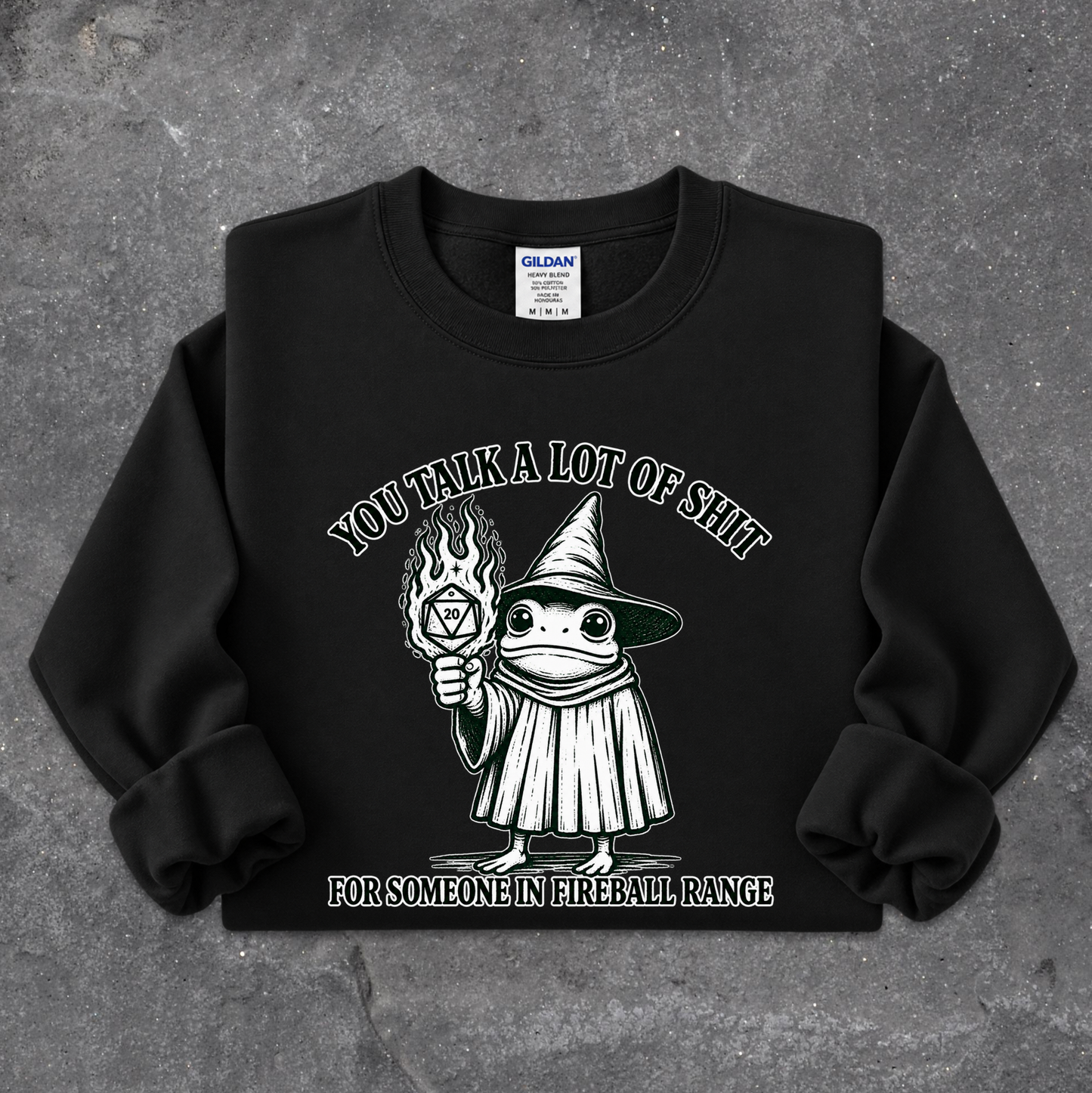 A black crewneck sweatshirt featuring a white graphic of a cartoon wizard holding a flaming torch, with the text 'YOU TALK A LOT OF SHIT FOR SOMEONE IN FIREBALL RANGE'. The sweatshirt is laid flat on a textured gray concrete surface, showing a Gildan brand tag inside the collar.