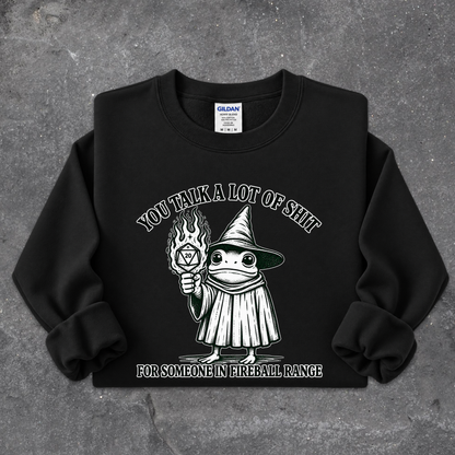 A black crewneck sweatshirt featuring a white graphic of a cartoon wizard holding a flaming torch, with the text 'YOU TALK A LOT OF SHIT FOR SOMEONE IN FIREBALL RANGE'. The sweatshirt is laid flat on a textured gray concrete surface, showing a Gildan brand tag inside the collar.