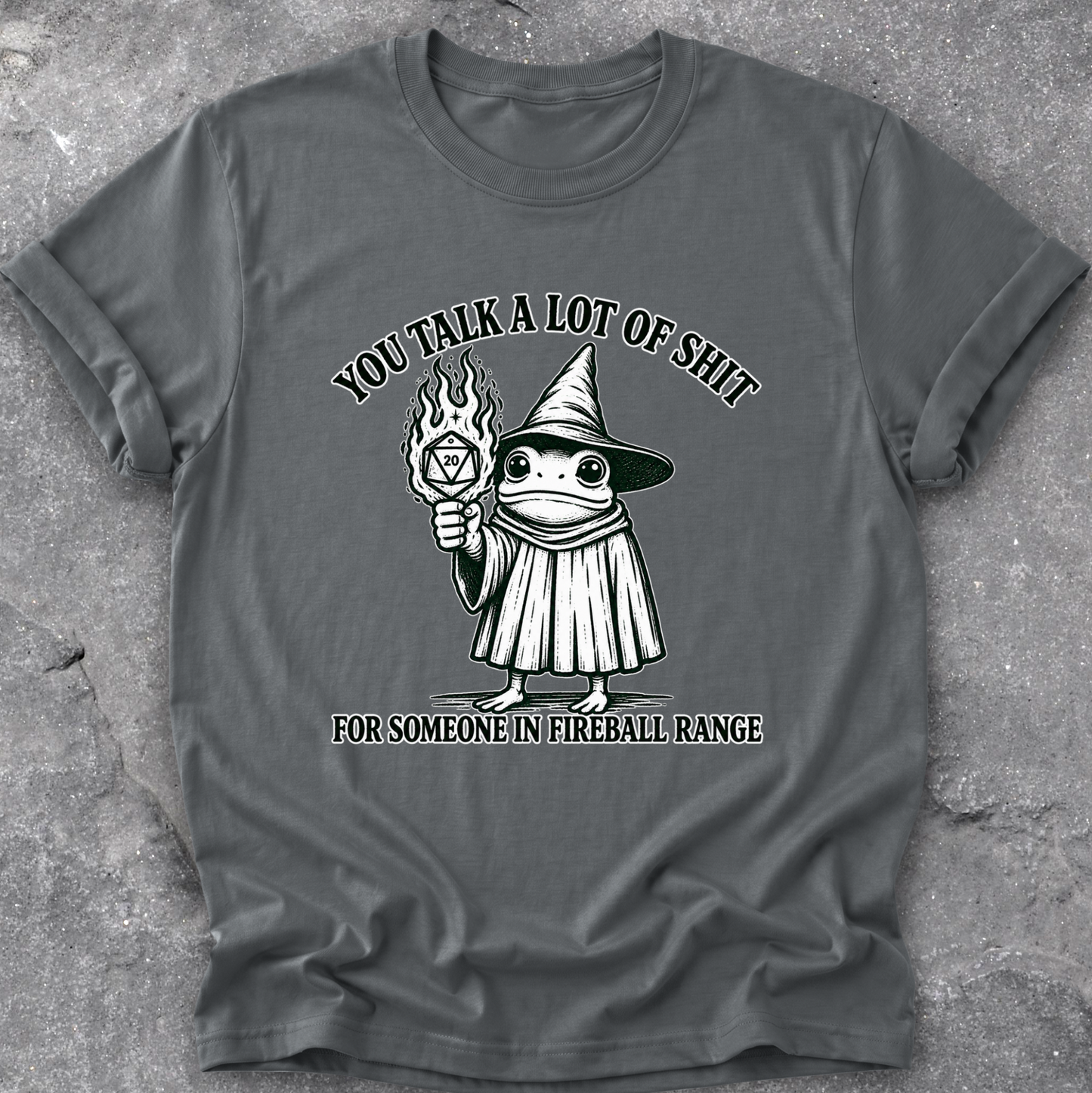 A dark gray crewneck t-shirt with a graphic of a cartoon frog in a wizard hat holding a fireball, with the text 'YOU TALK A LOT OF SHIT FOR SOMEONE IN FIREBALL RANGE'. The t-shirt is laid flat on a textured gray concrete surface.