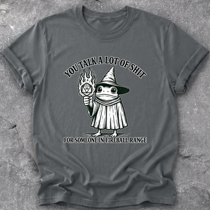 A dark gray crewneck t-shirt with a graphic of a cartoon frog in a wizard hat holding a fireball, with the text 'YOU TALK A LOT OF SHIT FOR SOMEONE IN FIREBALL RANGE'. The t-shirt is laid flat on a textured gray concrete surface.