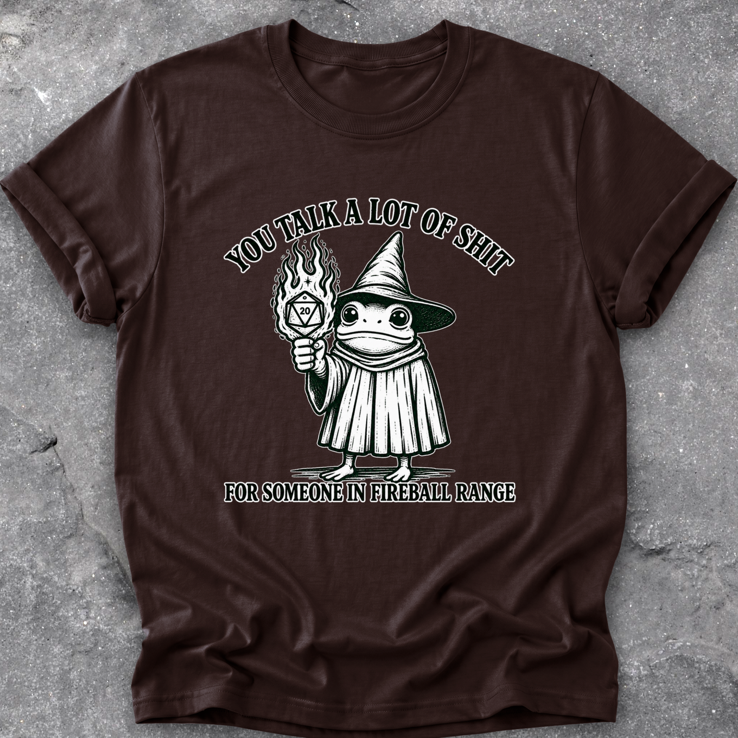 A dark brown short-sleeve t-shirt with a graphic of a cartoon frog in a wizard hat holding a fireball, with the text 'YOU TALK A LOT OF SHIT' above and 'FOR SOMEONE IN FIREBALL RANGE' below, laid flat on a gray concrete surface.