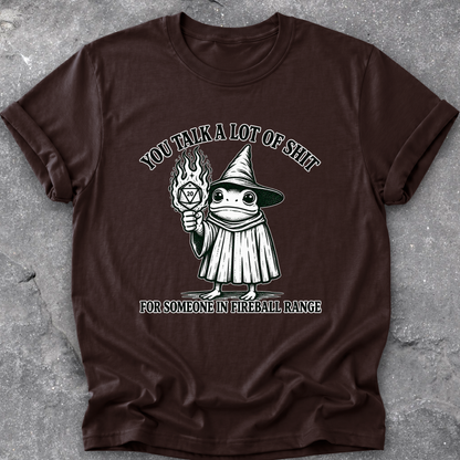 A dark brown short-sleeve t-shirt with a graphic of a cartoon frog in a wizard hat holding a fireball, with the text 'YOU TALK A LOT OF SHIT' above and 'FOR SOMEONE IN FIREBALL RANGE' below, laid flat on a gray concrete surface.