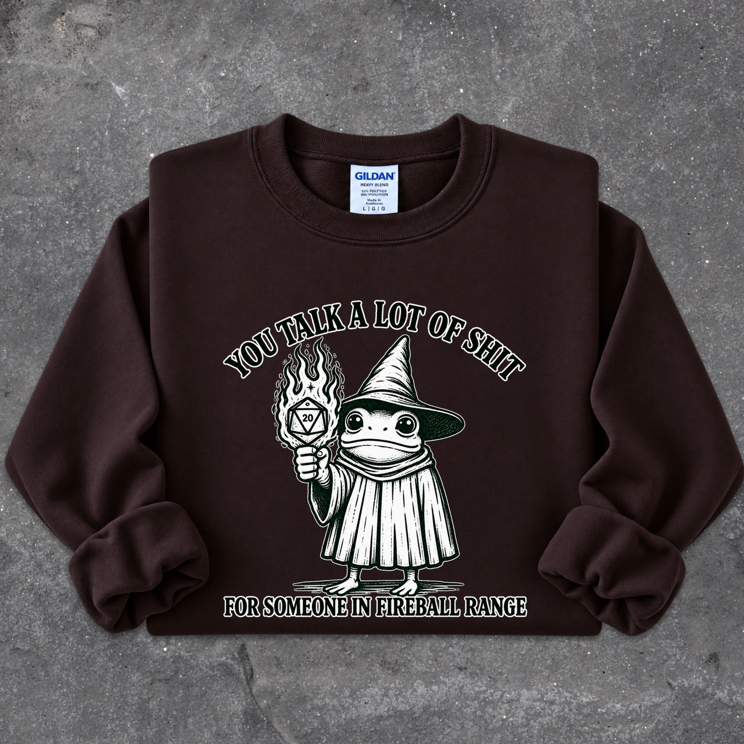 A dark brown crewneck sweatshirt laid flat on a textured gray background. The sweatshirt features a graphic of a cartoon witch holding a flaming torch, with the text 'YOU TALK A LOT OF SHIT' above and 'FOR SOMEONE IN FIREBALL RANGE' below.