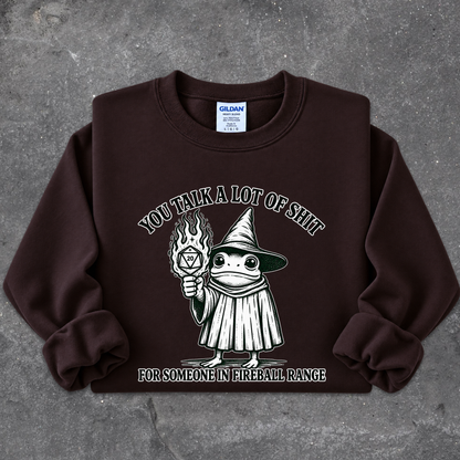 A dark brown crewneck sweatshirt laid flat on a textured gray background. The sweatshirt features a graphic of a cartoon witch holding a flaming torch, with the text 'YOU TALK A LOT OF SHIT' above and 'FOR SOMEONE IN FIREBALL RANGE' below.