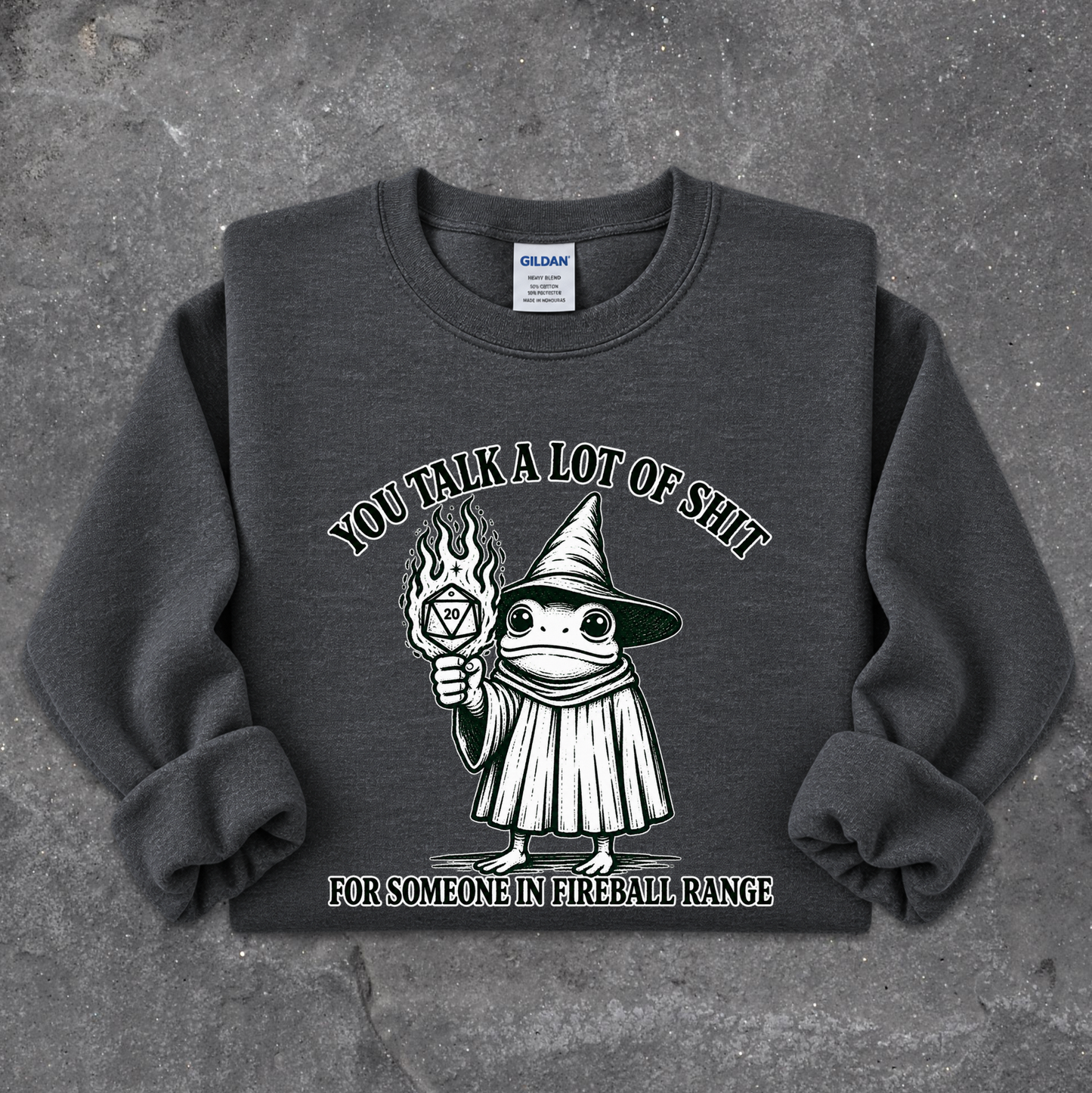 A dark gray crewneck sweatshirt featuring a cartoon graphic of a wizard holding a flaming orb, with the text 'YOU TALK A LOT OF SHIT' above and 'FOR SOMEONE IN FIREBALL RANGE' below, laid flat on a textured gray concrete surface.