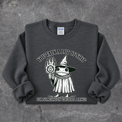 A dark gray crewneck sweatshirt featuring a cartoon graphic of a wizard holding a flaming orb, with the text 'YOU TALK A LOT OF SHIT' above and 'FOR SOMEONE IN FIREBALL RANGE' below, laid flat on a textured gray concrete surface.