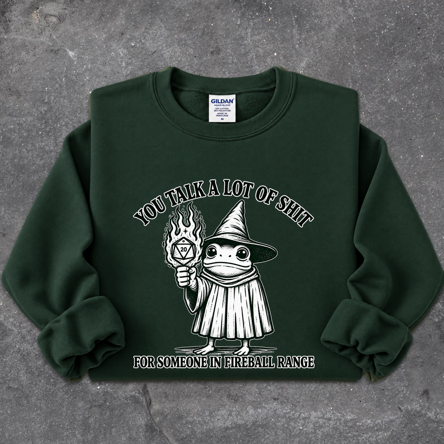 A dark green crewneck sweatshirt laid flat on a textured gray background. The sweatshirt features a white graphic of a cartoon frog dressed as a wizard holding a flaming torch, with the text 'YOU TALK A LOT OF SHIT' above and 'FOR SOMEONE IN FIREBALL RANGE' below.