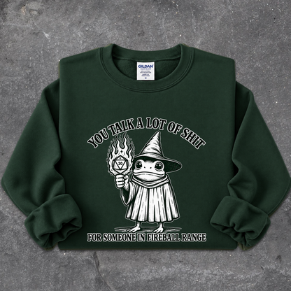A dark green crewneck sweatshirt laid flat on a textured gray background. The sweatshirt features a white graphic of a cartoon frog dressed as a wizard holding a flaming torch, with the text 'YOU TALK A LOT OF SHIT' above and 'FOR SOMEONE IN FIREBALL RANGE' below.