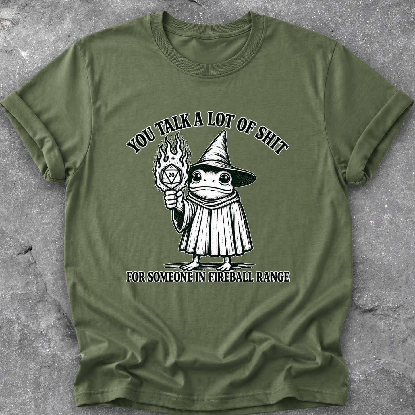 An olive green short-sleeve t-shirt with a black and white graphic of a wizard frog holding a fireball, with the text 'YOU TALK A LOT OF SHIT FOR SOMEONE IN FIREBALL RANGE', laid flat on a gray textured concrete surface.