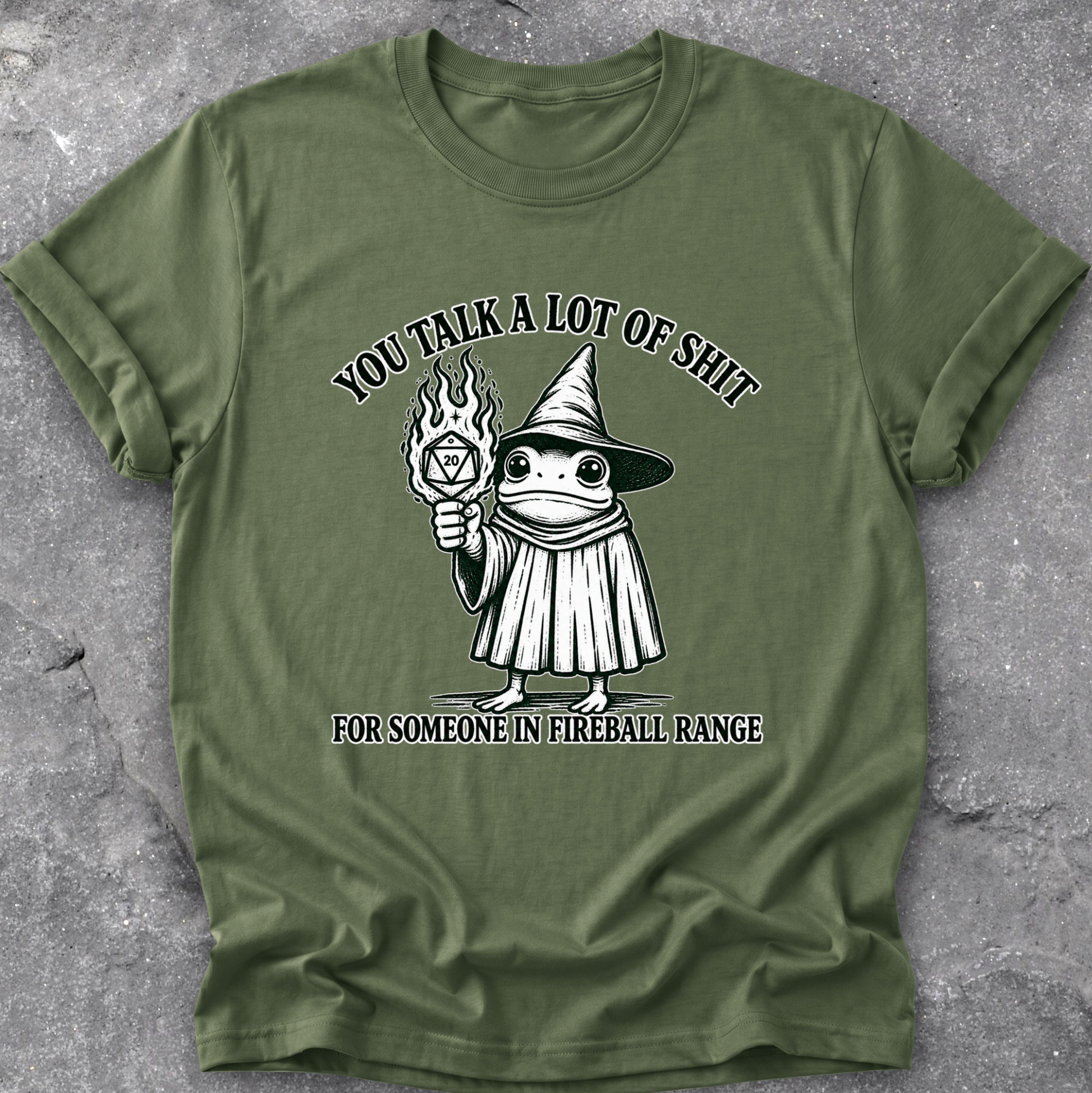 An olive green short-sleeve t-shirt with a black and white graphic of a wizard frog holding a fireball, with the text 'YOU TALK A LOT OF SHIT FOR SOMEONE IN FIREBALL RANGE', laid flat on a gray textured concrete surface.