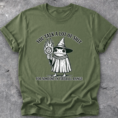 An olive green short-sleeve t-shirt with a black and white graphic of a wizard frog holding a fireball, with the text 'YOU TALK A LOT OF SHIT FOR SOMEONE IN FIREBALL RANGE', laid flat on a gray textured concrete surface.