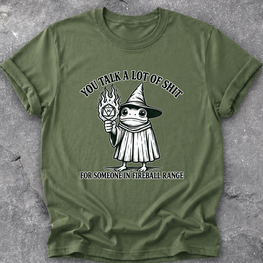 An olive green short-sleeve t-shirt with a black and white graphic of a wizard frog holding a fireball, with the text 'YOU TALK A LOT OF SHIT FOR SOMEONE IN FIREBALL RANGE', laid flat on a gray textured concrete surface.