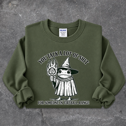 An olive green crewneck sweatshirt with a graphic of a cartoon fireball wizard holding a flame, with the text 'YOU TALK A LOT OF SHIT FOR SOMEONE IN FIREBALL RANGE'. The sweatshirt is laid flat on a textured gray concrete surface.