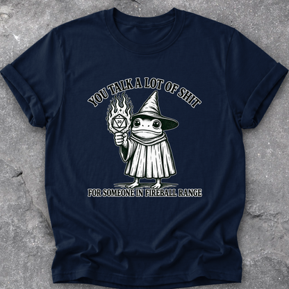 A navy blue short-sleeve t-shirt with a humorous graphic print, laid flat on a textured gray concrete surface. The graphic features a cartoon frog dressed as a wizard holding a fireball, with the text 'YOU TALK A LOT OF SHIT FOR SOMEONE IN FIREBALL RANGE'.
